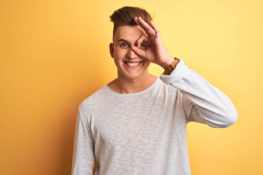 Beyaz rahat t-shirt giyen genç yakışıklı adam izole sarı arka plan üzerinde duran el gülümseyerek ok jest yapıyor, mutlu yüz ile parmaklardan bakan göz.