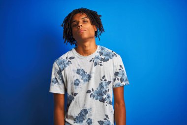 Yüzünde ciddi bir ifade ile izole mavi arka plan üzerinde yaz çiçek t-shirt giyen dreadlocks ile Afro Amerikan adam. Kameraya basit ve doğal bir bakış.