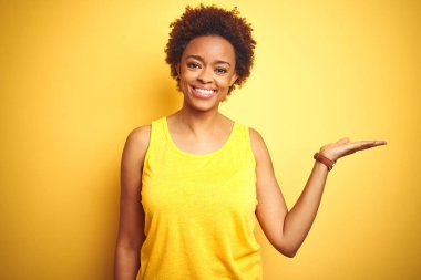 Beauitul afro-amerikan kadın izole sarı arka plan üzerinde yaz t-shirt giyen neşeli sunan gülümseyerek ve kameraya bakarak avuç içi ile işaret.