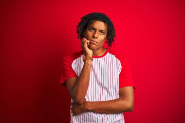 Dreadlocks çizgili t-shirt giyen Afro adam çene üzerinde el ile izole kırmızı arka plan üzerinde duran soru, dsive ifade düşünme. Düşünceli bir yüzle gülümsüyor. Şüphe kavramı.