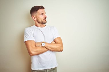 Genç yakışıklı adam izole arka plan üzerinde rahat beyaz t-shirt giyen kollarını ile yan bakarak ikna ve kendinden emin geçti