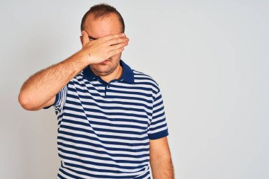Genç adam rahat çizgili polo giyen izole beyaz arka plan üzerinde el ile gözleri kaplayan ayakta, ciddi ve üzgün görünümlü. Görme, gizleme ve reddetme kavramı