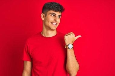 İzole kırmızı arka plan üzerinde duran t-shirt giyen genç hintli adam mutlu yüz görünümlü gülümseyerek ve başparmak ile yan işaret.