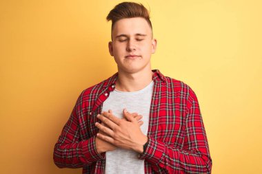 Genç yakışıklı adam kapalı gözleri ve yüzünde minnettar jest ile göğüs üzerinde gülümseyerek izole sarı arka plan üzerinde duran rahat gömlek giyiyor. Sağlık kavramı.