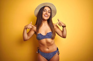 Genç güzel kadın tatilde bikini ve şapka giyen izole sarı arka plan üzerinde yüzünde gülümseme ile emin görünümlü, parmakları gururlu ve mutlu kendini işaret.