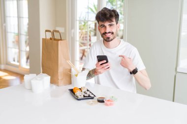 Genç adam eve teslim asya suşi yeme ve sürpriz yüz kendini işaret parmak ile akıllı telefon uygulaması kullanarak gıda sipariş