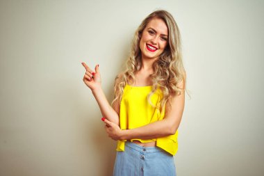 Yüzünde büyük bir gülümseme ile beyaz izole arka plan üzerinde duran sarı t-shirt giyen genç güzel kadın, kameraya bakarak yan el ve parmak ile işaret.