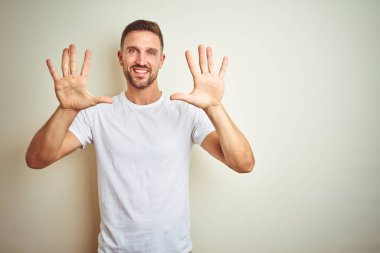 Gösterilen ve parmakları ile işaret izole arka plan üzerinde rahat beyaz t-shirt giyen genç yakışıklı adam kendinden emin ve mutlu gülümseyerek süre on numara.