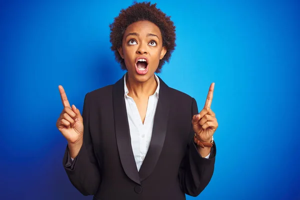 African american business executive woman over isolated blue background amazed and surprised looking up and pointing with fingers and raised arms.