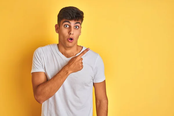 Young indian man wearing white t-shirt standing over isolated yellow background Surprised pointing with finger to the side, open mouth amazed expression.