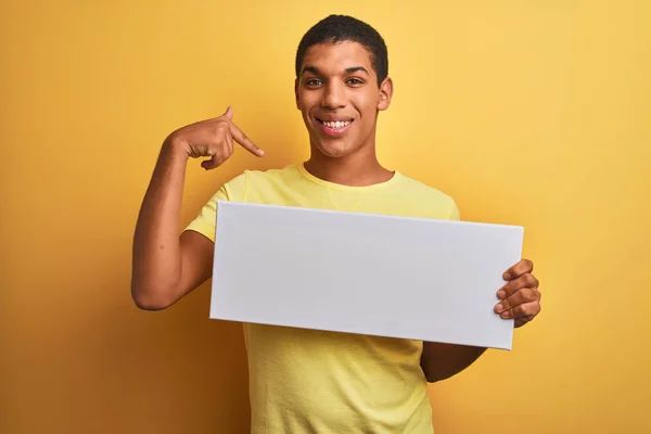 Young handsome arab man holding banner standing over isolated yellow background with surprise face pointing finger to himself