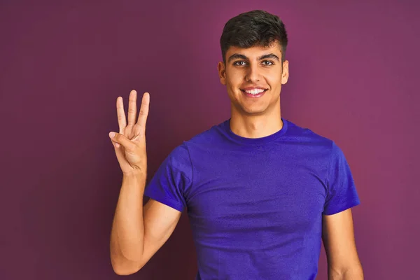 孤立した紫色の背景の上に立つTシャツを着た若いインド人男性が 自信を持って幸せに微笑みながら 3番の指を見せ 指を上げている — ストック写真
