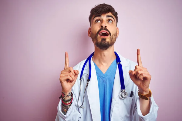 Young doctor man with tattoo wearing stethocope standing over isolated pink background amazed and surprised looking up and pointing with fingers and raised arms.
