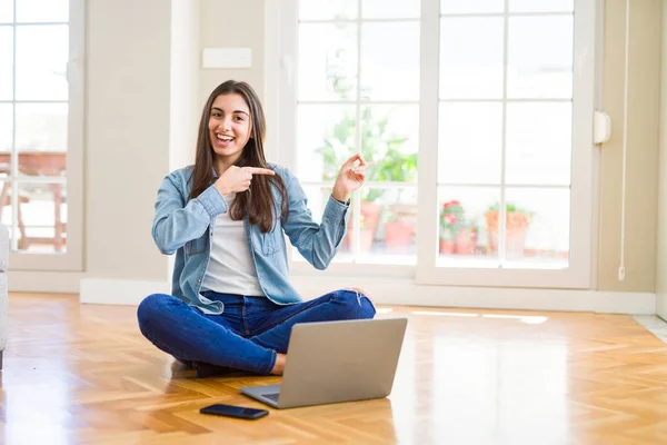 美丽的年轻女子坐在地板上 交叉的双腿使用笔记本电脑微笑 看着相机指着两只手和手指的一侧 — 图库照片
