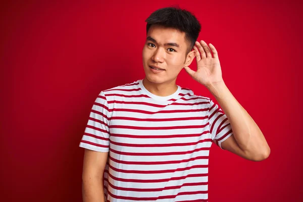 年轻的亚洲中国男子穿着条纹T恤站在孤立的红色背景微笑 手在耳朵上听听谣言或八卦 耳聋概念 — 图库照片