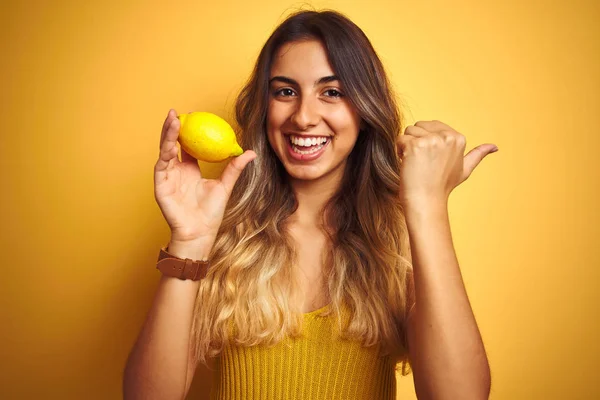 黄色の孤立した背景の上にレモンを食べて 幸せな顔で横に親指で示す若い美しい女性 — ストック写真
