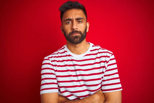Young indian man wearing striped t-shirt standing over isolated red background skeptic and nervous, disapproving expression on face with crossed arms. Negative person.