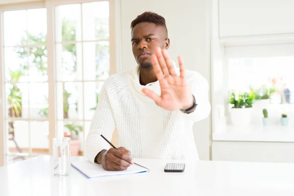 African american student man writing on a paper using a pencil with open hand doing stop sign with serious and confident expression, defense gesture