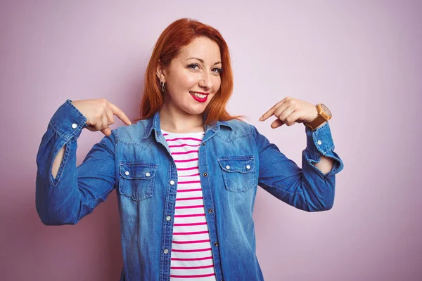 Denim gömlek giyen güzel kızıl saçlı kadın ve izole pembe arka plan üzerinde çizgili t-shirt yüzünde gülümseme ile emin görünümlü, parmakları gururlu ve mutlu kendini işaret.
