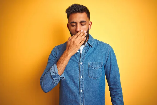 Young indian man wearing denim shirt standing over isolated yellow background bored yawning tired covering mouth with hand. Restless and sleepiness.