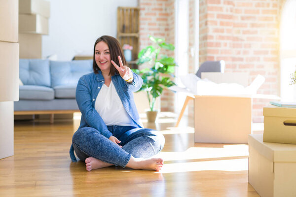 Young plus size woman sitting on the floor around cardboard boxes moving to a new home smiling with happy face winking at the camera doing victory sign. Number two.