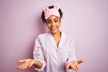 Genç afro-amerikan kadın izole pembe arka plan üzerinde pijama ve maske giyen neşeli sunan eller yardım ve kabul veren gülümseyerek.