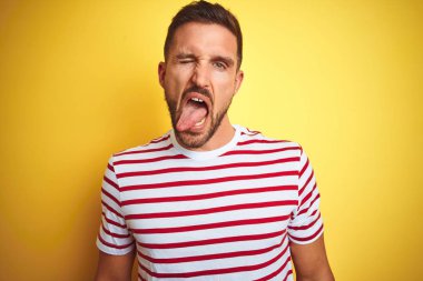 Genç yakışıklı adam sarı izole arka plan üzerinde rahat kırmızı çizgili t-shirt giyen komik ifade ile mutlu dil yapışmasını. Duygu kavramı.