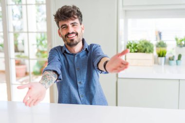 Beyaz masanın üzerinde oturan rahat gömlek giyen genç adam sarılmak için açık kollarla gülümseyerek kameraya bakıyor. Mutluluğu kucaklayan neşeli ifade.