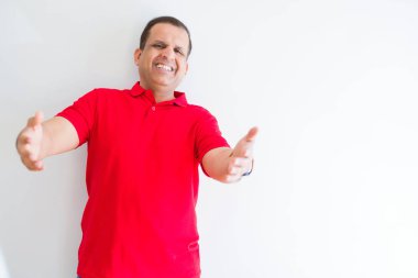 Beyaz duvarın üzerinde kırmızı tişört giyen orta yaşlı adam sarılmak için kollarını açarak gülümseyen kameraya bakıyor. Mutluluğu kucaklayan neşeli ifade.