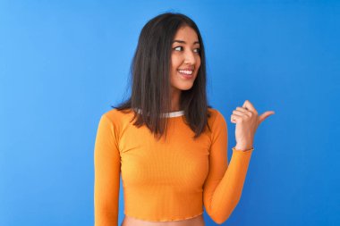 Turuncu t-shirt giyen genç güzel Çinli kadın izole mavi arka plan üzerinde duran mutlu yüz görünümlü ve başparmak ile yan işaret gülümseyerek.