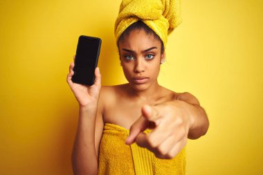 Duştan sonra havlu giyen Afro kadın kamera ve size parmak ile işaret izole sarı arka plan üzerinde smatrphone gösteren, el işareti, önden olumlu ve kendinden emin jest