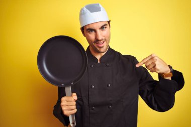 Üniforma ve şapka tutan aşçı tavası giyen genç şef, izole edilmiş sarı arka plan üzerinde sürpriz bir yüz le işaret eden parmakla kendine