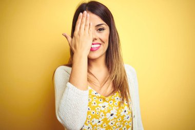 Bir gözünü eliyle kaplayan sarı izole arka plan üzerinde duran ceket giyen genç güzel kadın, yüzünde kendine güvenen bir gülümseme ve sürpriz duygu.