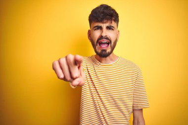 Çizgili t-shirt giyen dövme giyen genç adam izole sarı arka plan üzerinde duran hoşnut olmayan ve kameraya sinirli işaret, kızgın ve öfkeli size