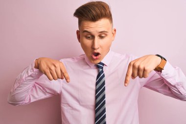 Genç yakışıklı işadamı gömlek ve kravat izole pembe arka plan üzerinde duran reklam gösteren parmakları ile aşağı işaret, sürpriz yüz ve açık ağız