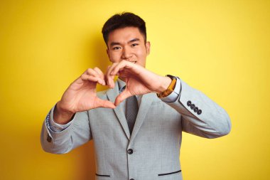 Takım elbise giyen ve kravat takan Asyalı çinli işadamı izole sarı arka plan üzerinde durarak sevgi kalp sembolü ve elleriyle şekil gösteren gülümseyerek. Romantik kavram.