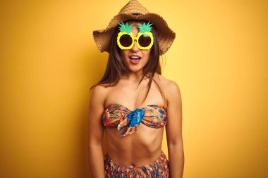 Bikini ve ananas güneş gözlüğü takan tatilde olan kadın, izole edilmiş sarı arka plan üzerinde korku ve sürpriz ifade, korku ve heyecanlı yüz ile şok.