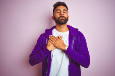 Mor sweatshirt giyen genç hintli adam kapalı gözleri ve yüzünde minnettar jest ile göğüs üzerinde gülümseyerek izole pembe arka plan üzerinde duran. Sağlık kavramı.