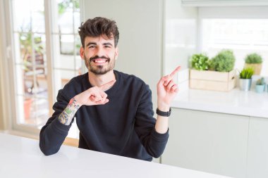 Beyaz masada oturan rahat sweatshirt giyen genç adam gülümseyerek ve yan iki el ve parmakları ile işaret kameraya bakıyor.