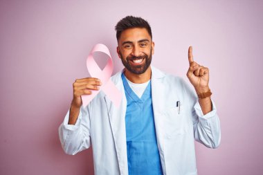 Genç Hint doktor adam izole pembe arka plan üzerinde duran kanser kurdele tutan mutlu yüz, bir numaralı ile bir fikir veya soru işaret parmağı ile şaşırttı