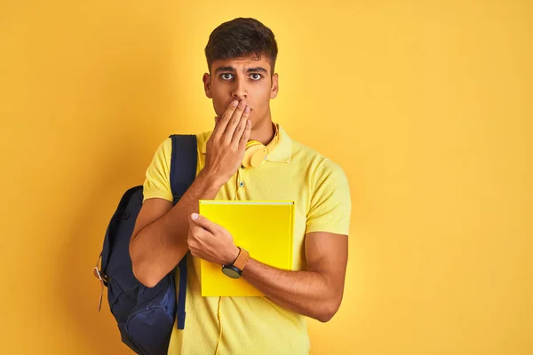 Indian student man wearing backpack headphones notebook over isolated yellow background cover mouth with hand shocked with shame for mistake, expression of fear, scared in silence, secret concept