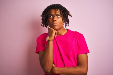 Afro amerikan adam dreadlocks t-shirt giyen izole pembe arka plan üzerinde çene üzerinde el soru, dsive ifade düşünme ile ayakta. Düşünceli bir yüzle gülümsüyor. Şüphe kavramı.