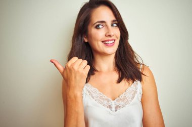 Beyaz izole arka plan üzerinde duran t-shirt giyen genç güzel kadın mutlu yüz görünümlü gülümseyerek ve başparmak ile yan işaret.