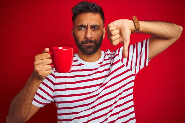 Young indian man wearing striped t-shirt drinking cup of coffee over isolated red background with angry face, negative sign showing dislike with thumbs down, rejection concept