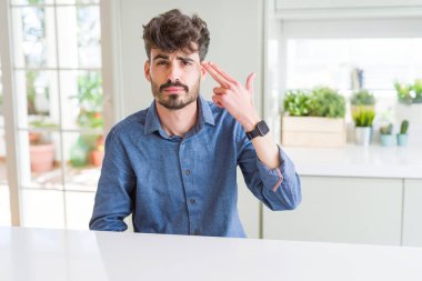 Beyaz masanın üzerinde oturan rahat gömlek giyen genç adam çekim ve kendini gösteren el ve parmaklarını kafaya, intihar jesti.