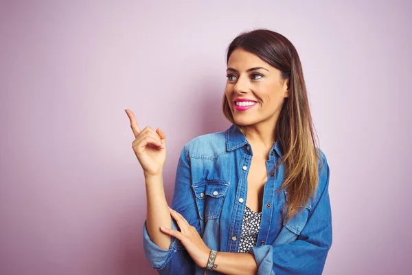 Young beautiful woman standing over pink isolated background with a big smile on face, pointing with hand and finger to the side looking at the camera.
