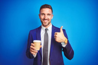 Genç yakışıklı iş adamı büyük bir gülümseme ok işareti yapıyor mutlu mavi izole arka plan üzerinde bir kağıt fincan üzerinde bir kahve içme, parmakları ile başparmak, mükemmel işareti