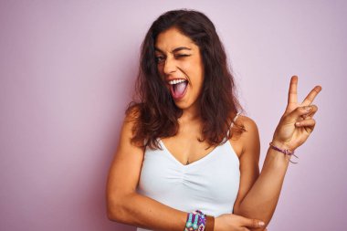 Beyaz tişört giyen genç güzel kadın izole pembe arka plan üzerinde duran mutlu yüz ile gülümseyerek zafer işareti yapıyor kameraya göz kırparak. İki numara.