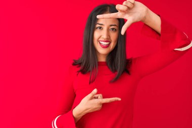 Genç güzel Çinli kadın rahat elbise giyen izole kırmızı arka plan üzerinde duran mutlu yüzü ile elleri ve parmakları ile çerçeve yapma gülümseyerek. Yaratıcılık ve fotoğrafçılık konsepti.