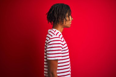 Yan bakan izole kırmızı arka plan üzerinde duran çizgili t-shirt giyen dreadlocks giyen genç afro adam, doğal yüz ve kendine güvenen gülümseme ile profil poz dinlenmek.